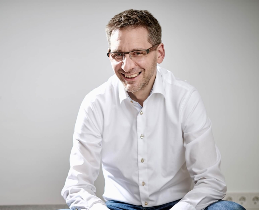 Das Profilbild von Henrik Zaborowski bei der Redneragentur PODIUM – Henrik Zaborowski sitzt entspannt auf einem Hocker, trägt ein weißes Hemd und lächelt in die Kamera.