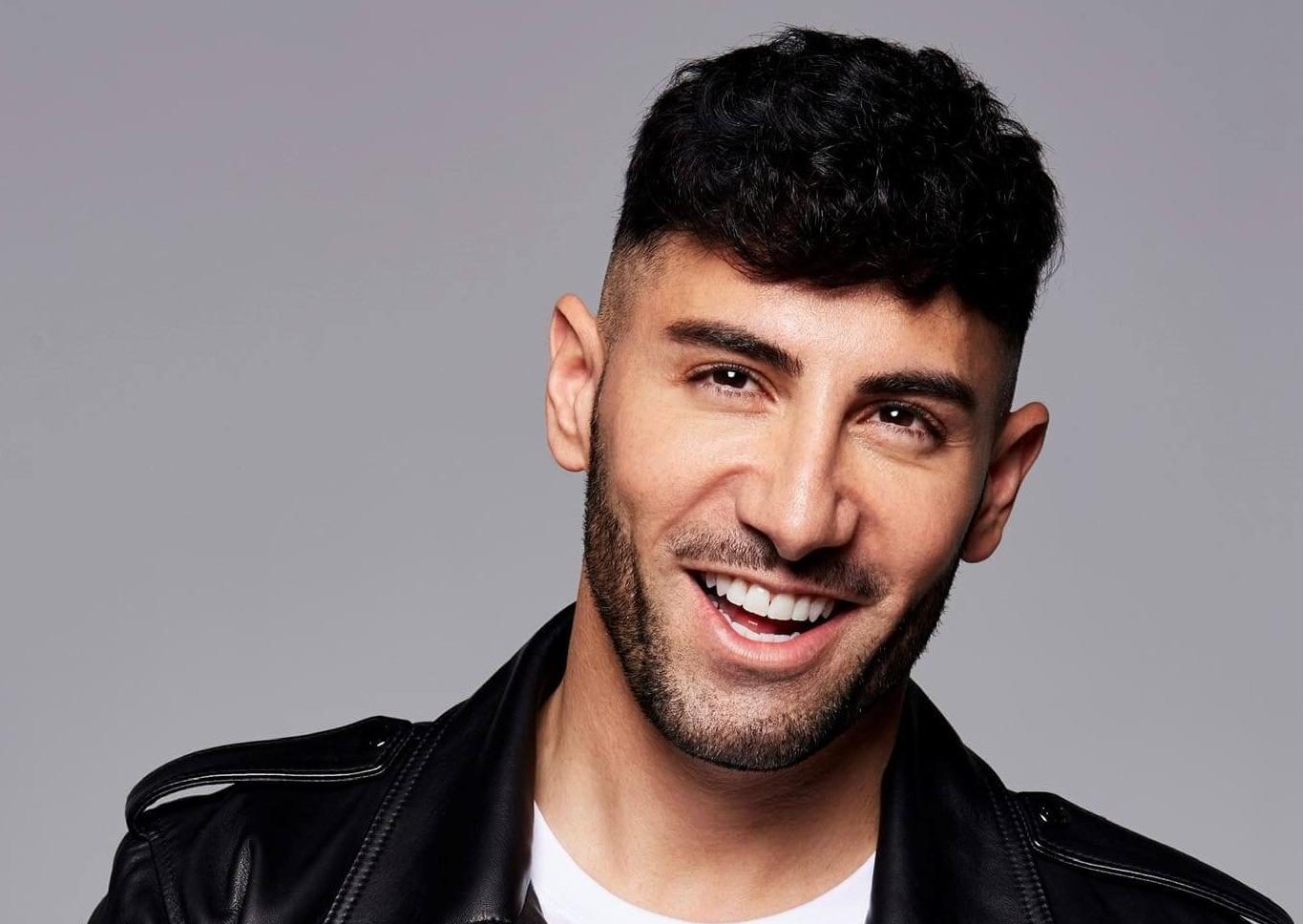 Das Profilbild von Ayan Yaruk bei der Redneragentur PODIUM – Ayan Yaruk lächelt in die Kamera, trägt eine schwarze Lederjacke und weißes Shirt vor grauem Hintergrund.