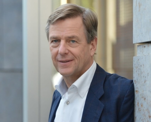 Dr. Claus Kleber, bekannter deutscher Journalist und Redner, steht in einem modernen urbanen Umfeld und trägt ein dunkelblaues Sakko über einem weißen Hemd.