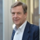 Dr. Claus Kleber, bekannter deutscher Journalist und Redner, steht in einem modernen urbanen Umfeld und trägt ein dunkelblaues Sakko über einem weißen Hemd.