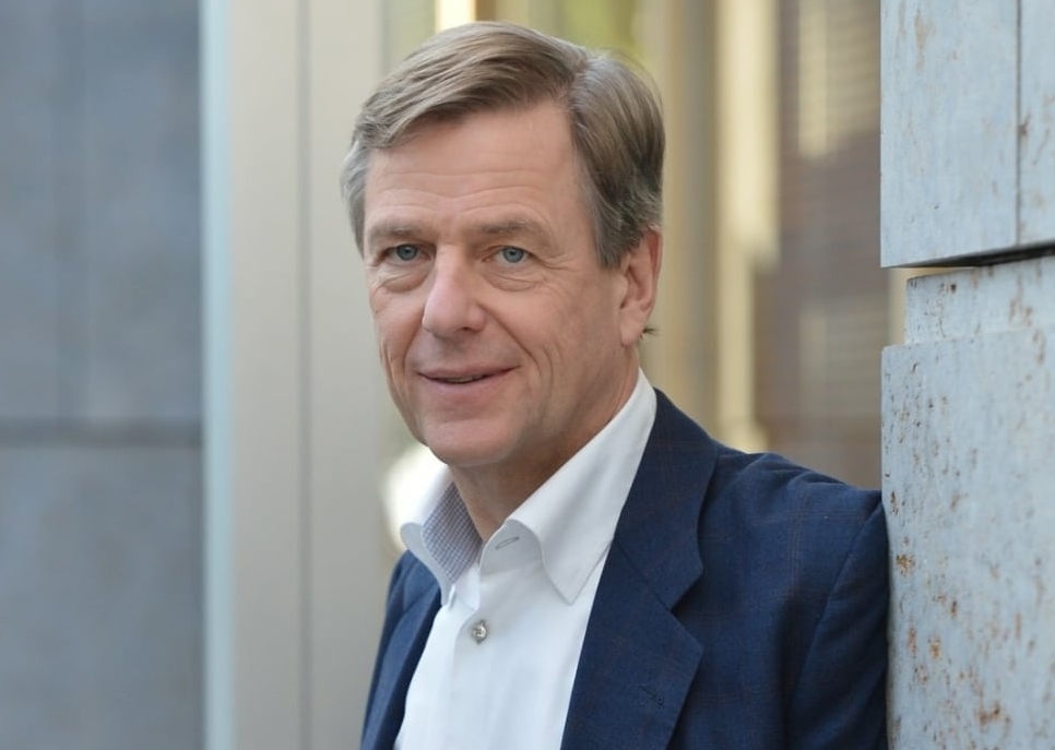 Dr. Claus Kleber, bekannter deutscher Journalist und Redner, steht in einem modernen urbanen Umfeld und trägt ein dunkelblaues Sakko über einem weißen Hemd.