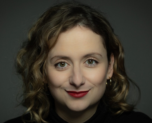 Das Profilbild von Alexandra Zykunov bei der Redneragentur PODIUM – Alexandra Zykunov mit lockigem Haar, rotem Lippenstift und direktem Blick in die Kamera vor dunklem Hintergrund.