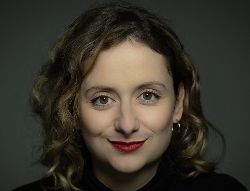 Das Profilbild von Alexandra Zykunov bei der Redneragentur PODIUM – Alexandra Zykunov mit lockigem Haar, rotem Lippenstift und direktem Blick in die Kamera vor dunklem Hintergrund.