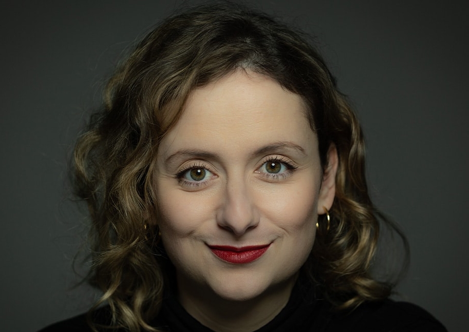 Das Profilbild von Alexandra Zykunov bei der Redneragentur PODIUM – Alexandra Zykunov mit lockigem Haar, rotem Lippenstift und direktem Blick in die Kamera vor dunklem Hintergrund.