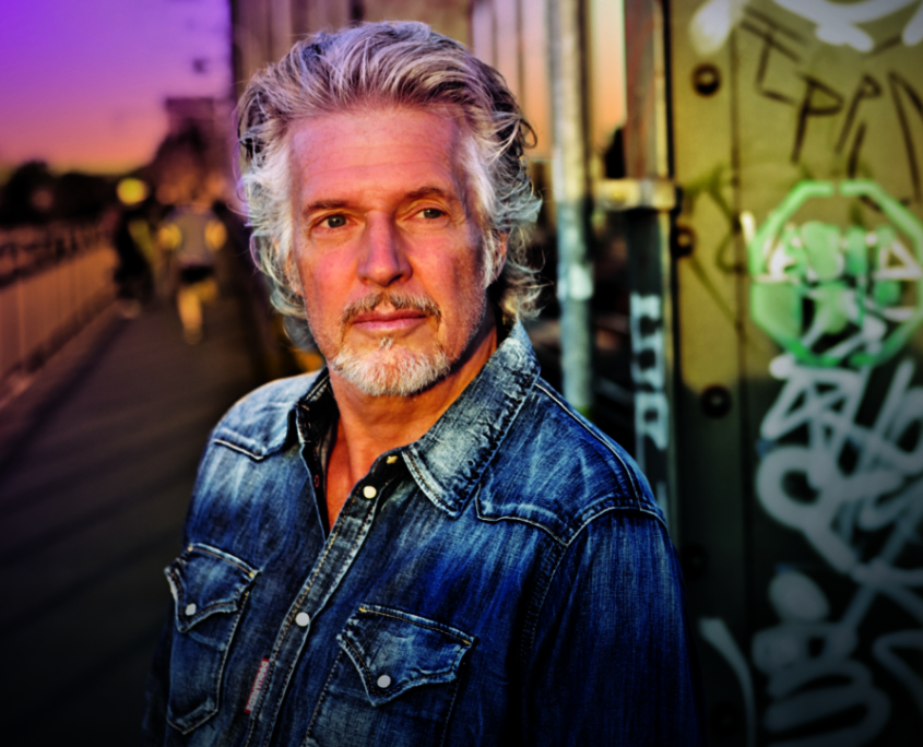 Das Profilbild von Frank Schätzing bei der Redneragentur PODIUM zeigt ihn vor einer urbanen Kulisse mit Graffiti und Sonnenuntergang – ein ausdrucksstarkes Porträt des bekannten Redners.