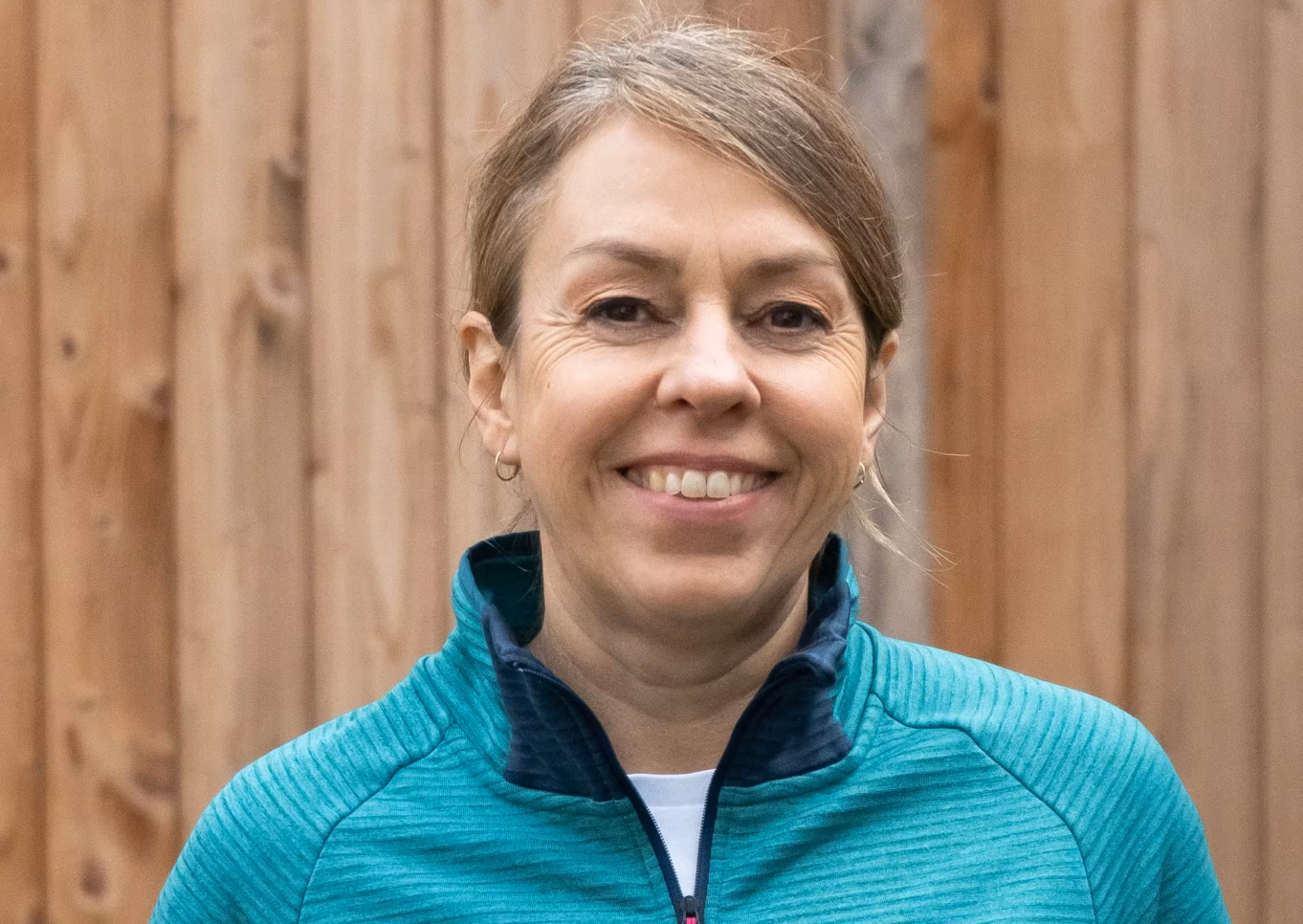Das Profilbild von Melanie Bleicher bei der Redneragentur PODIUM vor einem Holz-Hintergrund, mit freundlichem Gesichtsausdruck und sportlicher Kleidung.