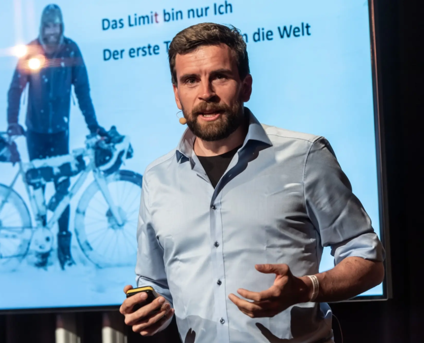 Das Profilbild von Jonas Deichmann bei der Redneragentur PODIUM – als Redner auf der Bühne mit Präsentation im Hintergrund