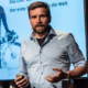 Das Profilbild von Jonas Deichmann bei der Redneragentur PODIUM – als Redner auf der Bühne mit Präsentation im Hintergrund