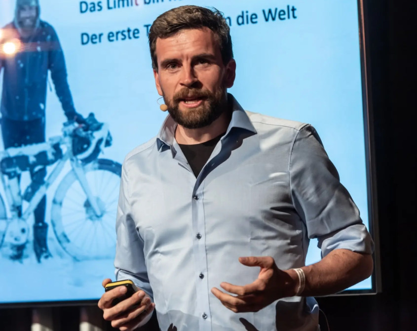 Das Profilbild von Jonas Deichmann bei der Redneragentur PODIUM – als Redner auf der Bühne mit Präsentation im Hintergrund