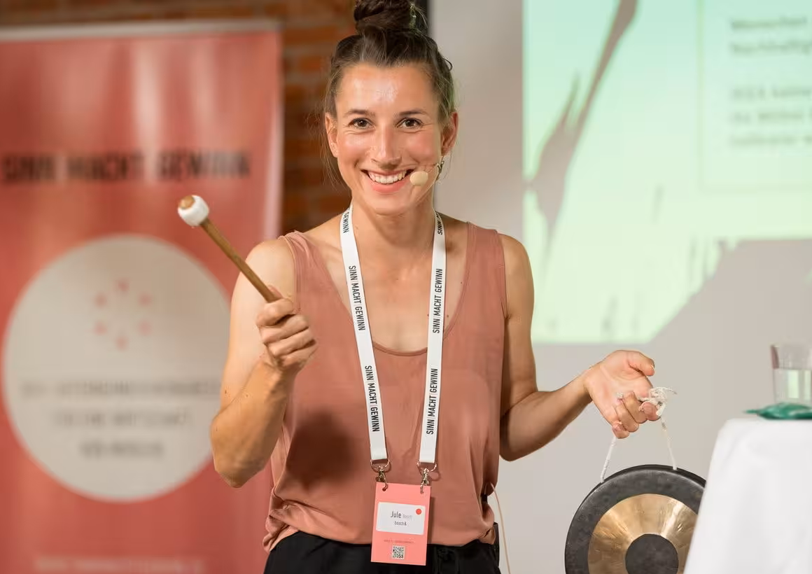Das Profilbild von Jule Bosch bei der Redneragentur PODIUM – Jule Bosch hält einen Gong und spricht auf einer Bühne.