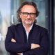 Peter Brandl im Business-Outfit mit verschränkten Armen vor einem modernen Glasgebäude – das Profilbild bei der Redneragentur PODIUM