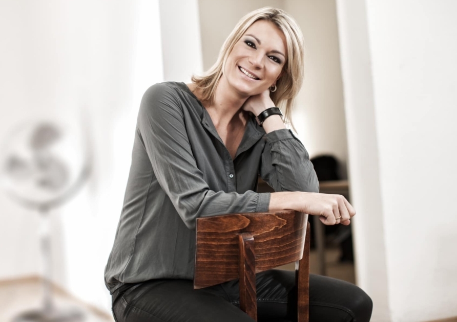 Maria Höfl-Riesch lächelt entspannt, sitzend auf einem Stuhl in einem hellen Raum.